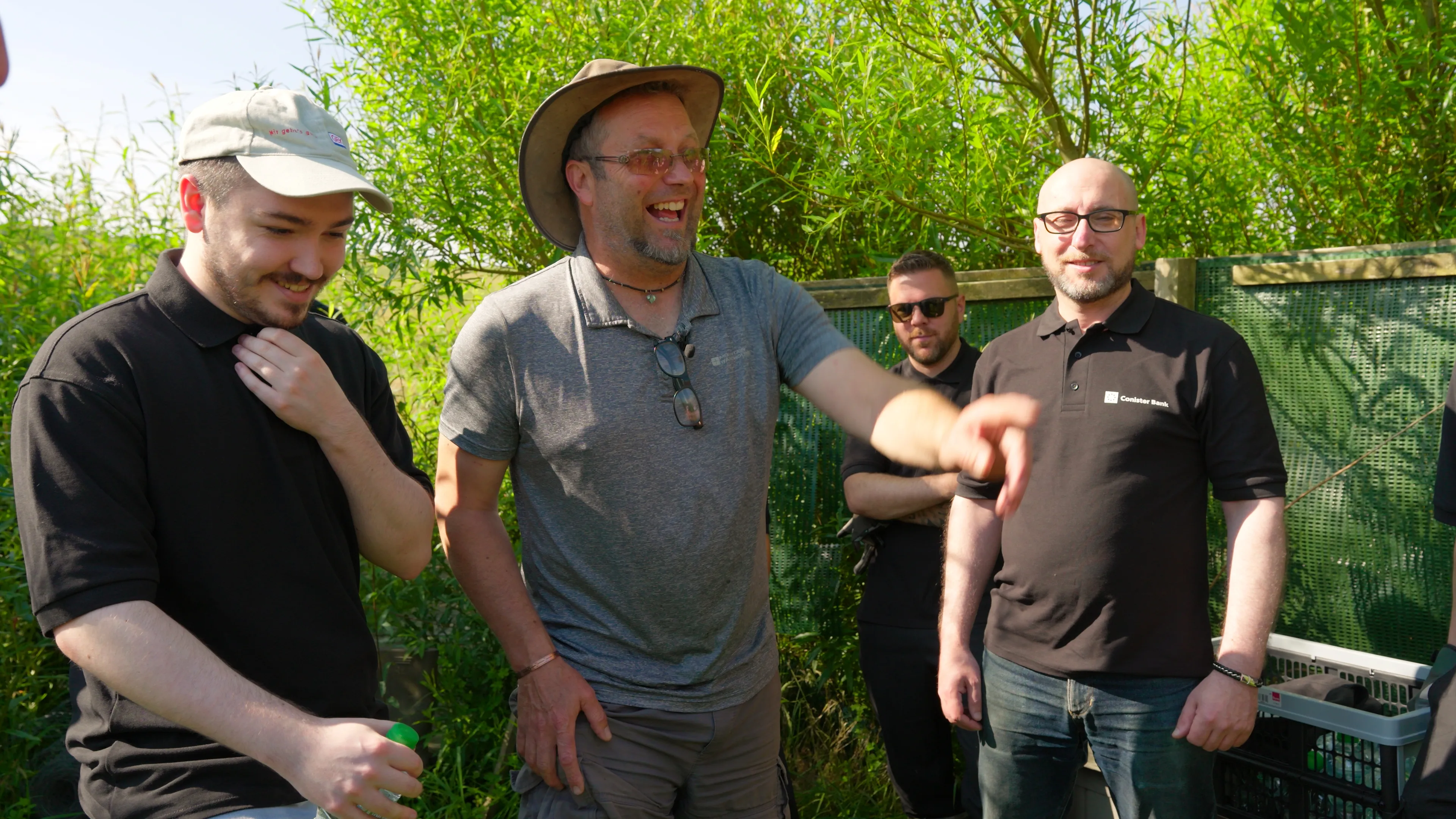 A group of five men stands outdoors, smiling and chatting. One man in the center wears a wide-brimmed hat, gesturing energetically. The mood is lively and relaxed.